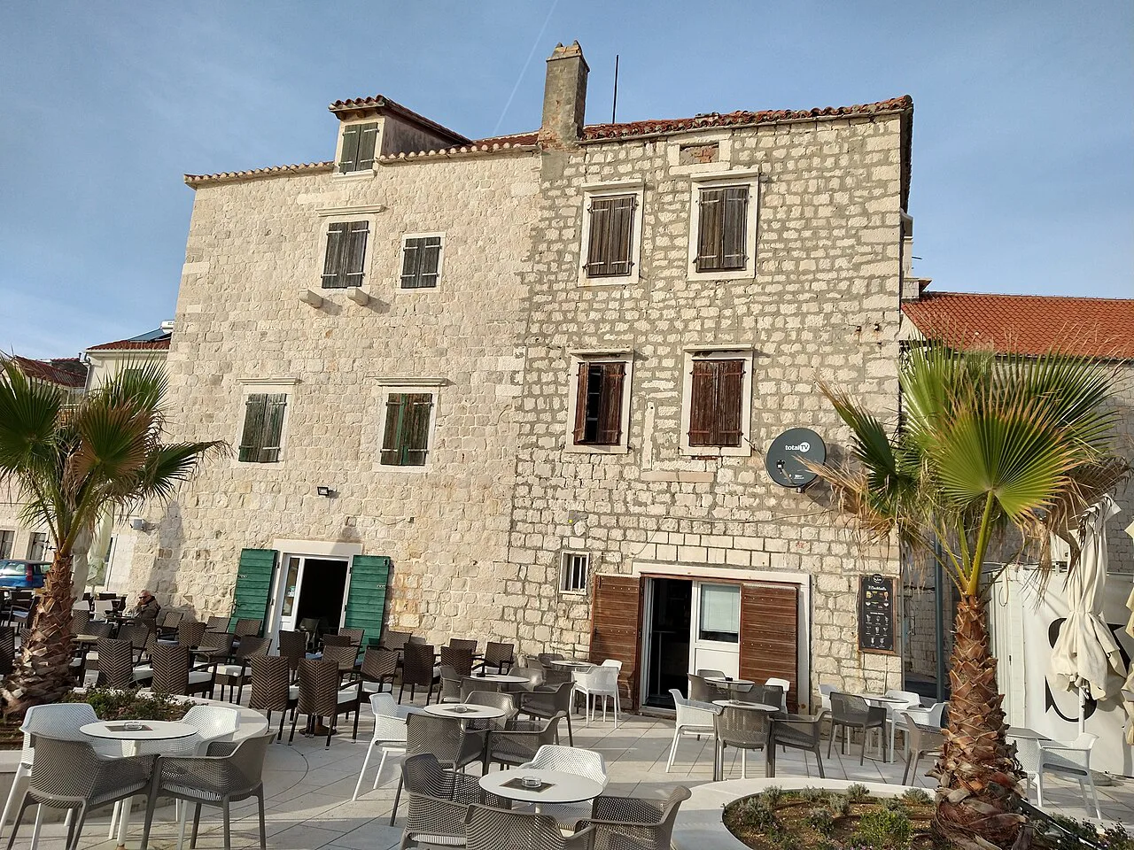 A stone building with a café terrace and palm trees in Kaštel Stari, Dalmatia