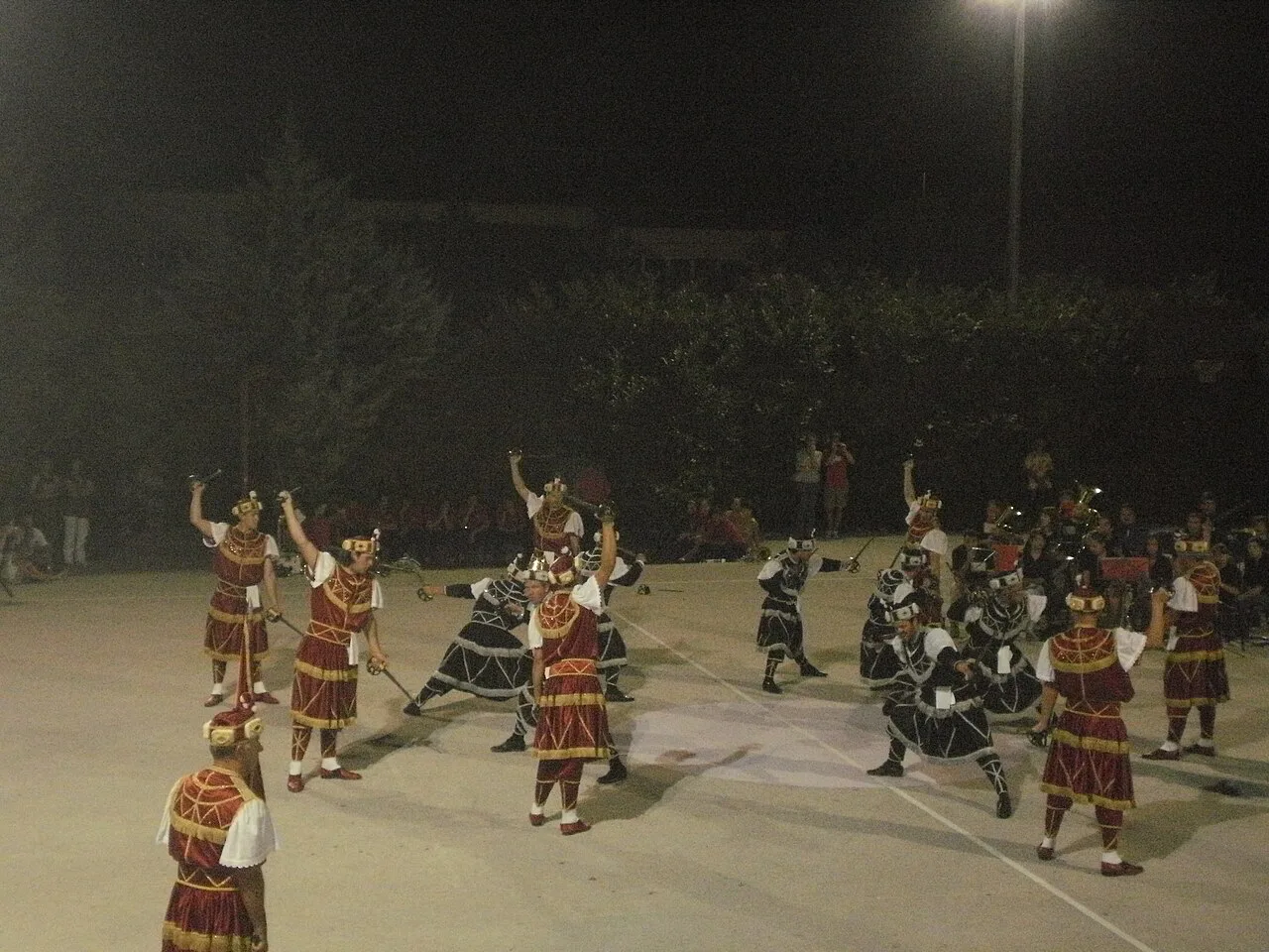 Dancers in red and black costumes performing the Moreška sword dance on an open-air stage.