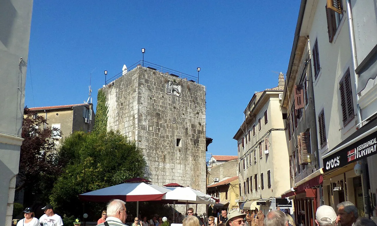 A medieval stone tower beside café umbrellas, seen from Liberty Square in Poreč
