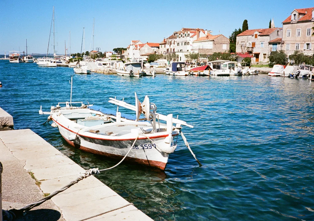 A small wooden boat moored at the stone quay in Zlarin, with waterfront houses behind, Šibenik